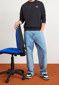 Svart sweatshirt med logotyp, ljusblå baggyjeans med lappar, svartvita sneakers, stående bredvid en blå och svart kontorsstol.