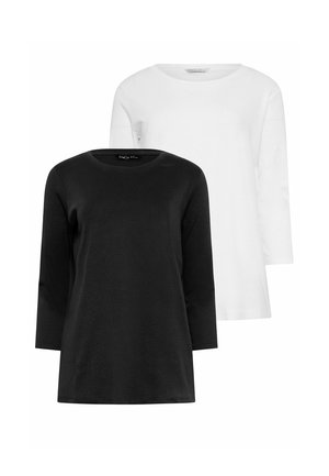 Eine Frau steht vor einer weißen Wand und trägt ein schwarzes Langarmshirt, hochgekrempelte blaue Jeans und weiße Turnschuhe, mit einer Hand in der Tasche.