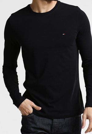 Mand iført en ensfarvet sort langærmet trøje med en lille Tommy Hilfiger-logo på venstre bryst, hånd i lommen.