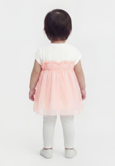 Bambino in piedi di spalle, indossa una maglietta bianca con un soprabito in tulle rosa, leggings bianchi e scarpe bianche su uno sfondo semplice.