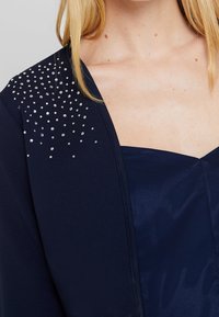 Mörkblå jacka med en slät textur, som har en dekorativ samling av små rhinestones på en axel.