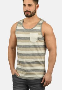 Camiseta sin mangas para hombre en beige claro con rayas horizontales en gris y gris oscuro. Presenta un pequeño bolsillo en el pecho izquierdo y una textura de tela suave.