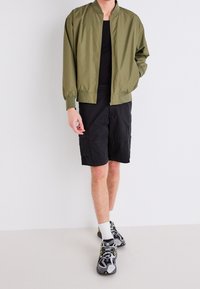 Giacca bomber verde oliva con polsini elasticizzati, pantaloni cargo neri e scarpe sportive grigie con accenti gialli, indossata con calzini bianchi.