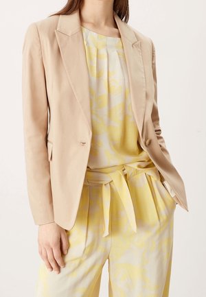 Mujer con blazer beige sobre blusa floral amarilla y blanca y pantalones de pierna ancha a juego con cinturón de lazo, con la mano en el bolsillo.