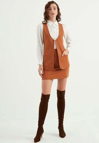 Brun skräddarsydd väst över en vit skjorta, tillsammans med en brun minikjol. Modellen bär bruna knähöga stövlar som visar en elegant design.
