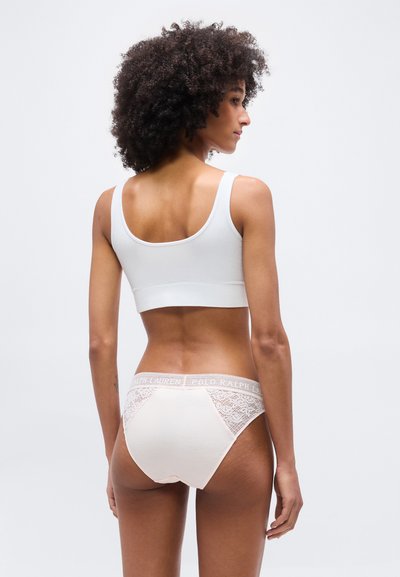 Γυναίκα με σγουρά μαλλιά στέκεται με την πλάτη γυρισμένη, φορώντας άσπρο κοντό τοπ και μπεζ εσώρουχο με δαντέλα και λευκή ζώνη μέσης «Polo Ralph Lauren».