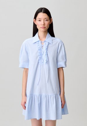 Donna con capelli lunghi e scuri indossa un vestito azzurro chiaro a maniche corte con colletto, pizzi arricciati sul davanti e orlo raccolto, in piedi davanti a uno sfondo semplice.