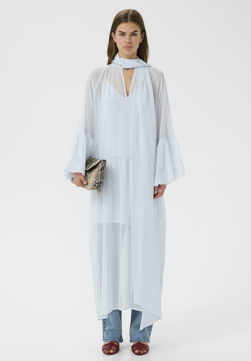 Femme portant une robe légère bleu clair transparente par-dessus un jean, tenant une pochette en peau de serpent, avec des chaussures ouvertes tressées marron et une écharpe assortie.