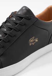 Gros plan sur une basket en cuir noir avec des lacets noirs, une semelle blanche, un intérieur beige et un logo crocodile doré sur le côté.