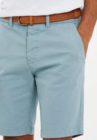 Lyseblå shorts lavet af vævet stof, med flad front, sidelommer og et brunt flettet bælte med spænde.