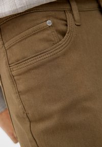 Brązowe spodnie z mieszanki bawełny o gładkiej fakturze, z przednią kieszenią i srebrnym detalem w postaci nitu. Widoczne akcenty przeszyć i szwów.