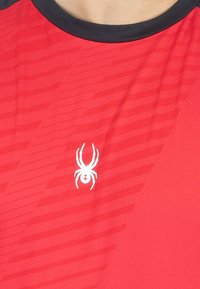 Camiseta atlética roja con cuello negro, que presenta un logotipo de araña blanca y un sutil patrón de rayas diagonales en la tela.