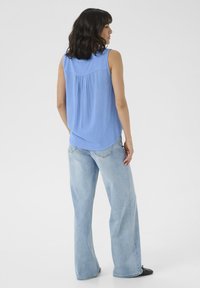 Top sans manches bleu clair avec un dos plissé, associé à un jean large délavé, sur fond blanc uni.