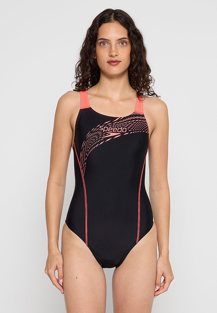 Donna con capelli ricci e scuri che indossa un costume da bagno intero Speedo nero con spalline corallo e un design grafico rosa sul davanti, in piedi contro uno sfondo bianco.