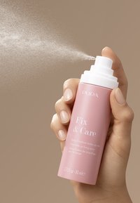 Różowa cylindryczna butelka ze spryskiwaczem i białą pompką, oznaczona napisem "Fix & Care", rozpylająca delikatną mgiełkę na neutralnym tle.