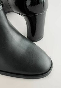 Botine din piele neagră până la gleznă au o finisare netedă și texturată și un toc înalt negru, strălucitor. Designul include un vârf rotunjit și cusături întărite.