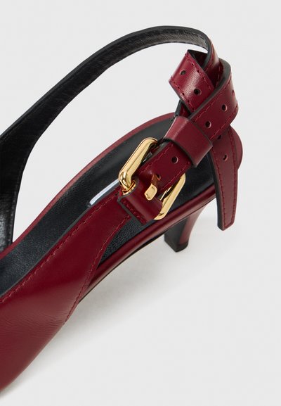 Red leather slingback shoe with a black interior, featuring a gold buckle and perforated accents, on a small black heel.