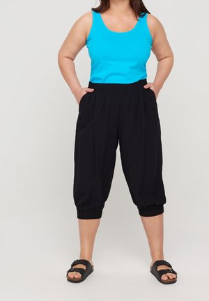 Frau trägt ein leuchtend blaues ärmelloses Oberteil, schwarze verkürzte Hosen mit elastischen Bündchen und schwarze Sandalen mit offenen Zehen, steht mit den Händen in den Taschen.