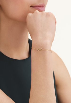 Vrouw draagt een delicaat gouden armbandje met een oneindigheidssymbool en kleine steentjes om haar pols, gekleed in een mouwloze zwarte top.