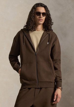Ung mand med langt krøllet hår, iført mørke solbriller, brun lynlås-hættetrøje, beige skjorte, matchende brune bukser og et armbåndsur.
