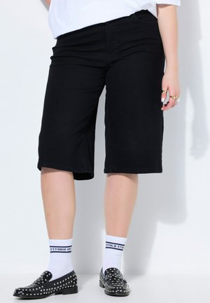 Person iført sorte knælange shorts, hvide sokker med sort tekst og sorte loafers med nitter, står op ad en hvid væg.