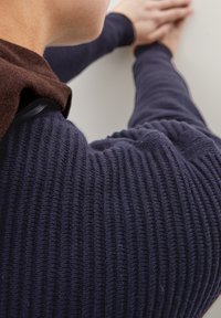 Marineblauer gerippter Pullover mit einem weichen, braunen Fleecepulloverkragen. Nahaufnahme des texturierten Stoffs, der das gestapelte Design und die nahtlose Verarbeitung zeigt.