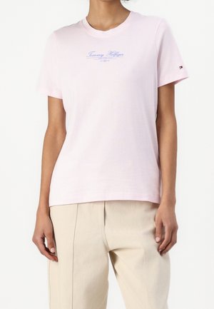 Vrouw met een lichtroze Tommy Hilfiger T-shirt met blauwe logotekst en beige broek tegen een effen witte achtergrond.