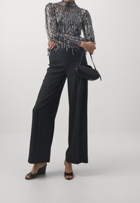 Schwarzes, langärmeliges Oberteil mit Pailletten, kombiniert mit weiten schwarzen Hosen und offenen Keilsandalen. Inklusive kleiner schwarzer Handtasche.