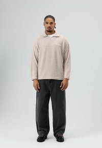 Beige fleece trui met een kraag en lange mouwen, voorzien van een klein logo aan de linkerkant; gecombineerd met zwart losvallende jeans en zwarte schoenen.
