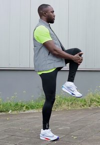 Idrottare i en grå väst över en neongrön tröja, svarta tights och vita sportskor, som utför en knälyft utomhus.