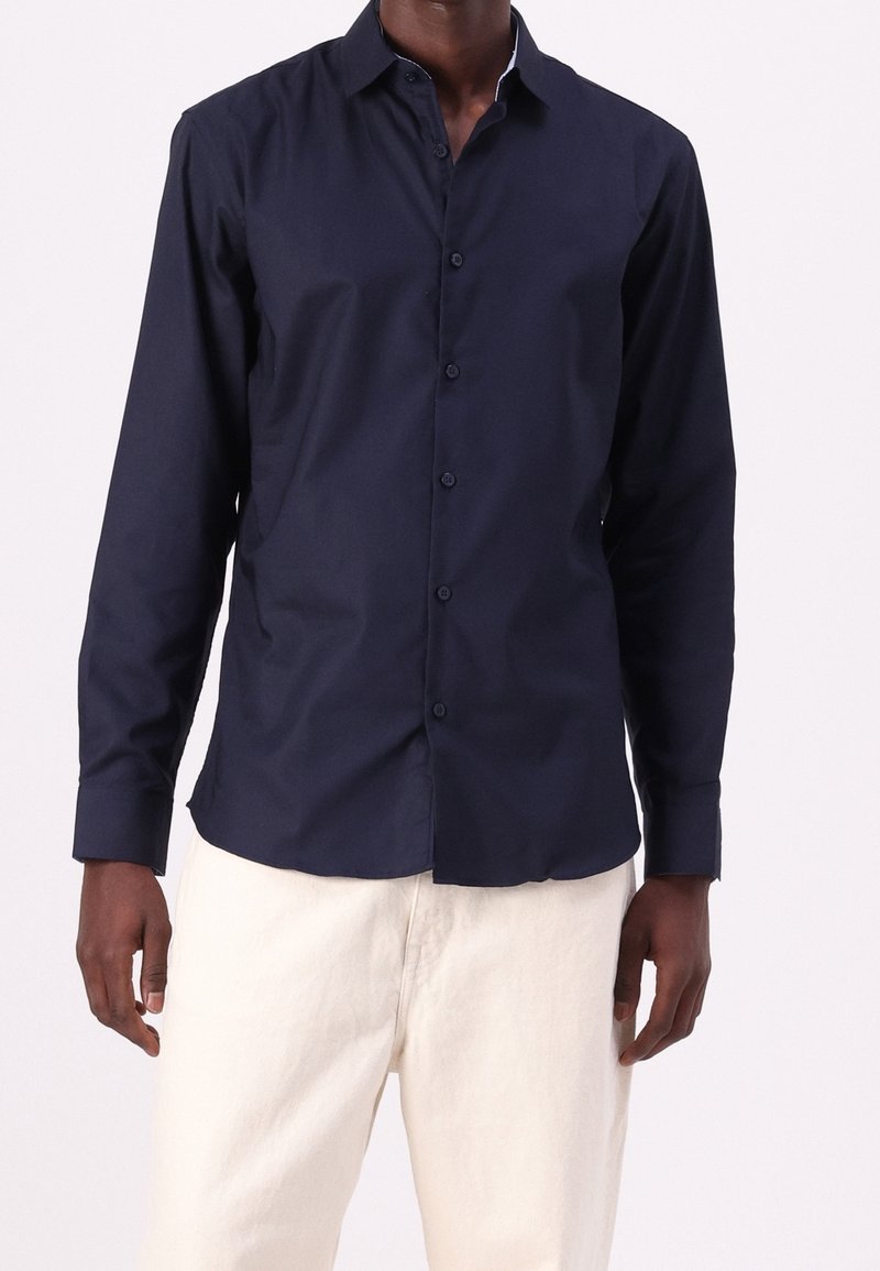 Homme portant une chemise ajustée bleu marine foncé à manches longues boutonnée et un pantalon beige clair sur un fond uni.