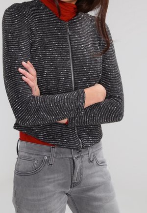 Černá a stříbrná pruhovaná bunda na zip s dlouhými rukávy a přiléhavým střihem, přes červený rolák. Šedé džíny viditelné dole.