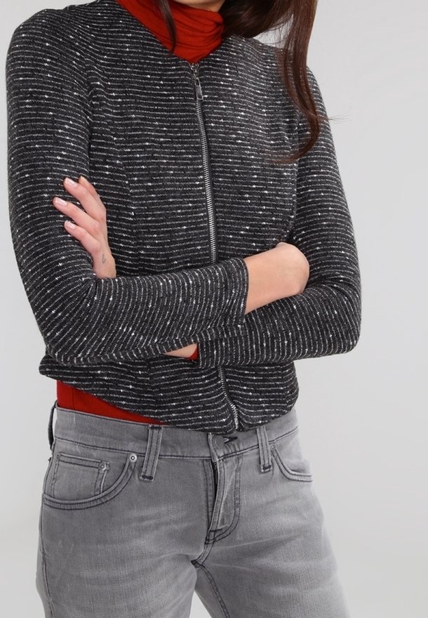 Kurtka zapinana na zamek błyskawiczny w czarno-srebrne paski, z długimi rękawami i dopasowanym krojem, nałożona na czerwony golf. Widoczne szare dżinsy poniżej.