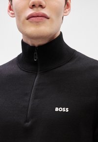 Camisola preta com fecho, com um colarinho alto, textura canelada, apresentando um logotipo branco "BOSS" no peito. Tecido elástico e ajuste justo.