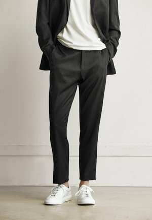 Personne portant un pantalon noir sur mesure, un blazer noir, un t-shirt blanc et des baskets blanches, debout les mains dans les poches devant un fond uni.