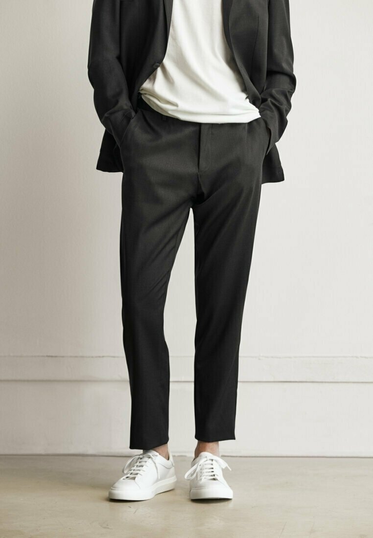 Personne portant un pantalon noir sur mesure, un blazer noir, un t-shirt blanc et des baskets blanches, debout les mains dans les poches devant un fond uni.