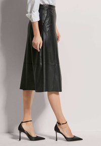 Schwarzer Lederrock mit ausgestelltem Design, seitlichen Nähten und hoher Taille. Kombiniert mit schwarzen spitzen High Heels und Knöchelriemen.