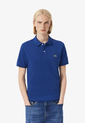 Persona con capelli biondi di lunghezza spalla che indossa una polo blu con il logo verde Lacoste, in piedi davanti a uno sfondo bianco.