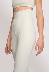 Leggings a vita alta di colore crema realizzati in tessuto elasticizzato, dotati di tasche laterali e una superficie liscia e opaca. I dettagli delle cuciture sono visibili.
