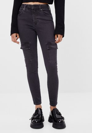 Frau trägt schwarze High-Waist Skinny Jeans mit Cargotaschen und schwarze klobige Schuhe mit Schnalle, steht vor einem einfarbigen Hintergrund.