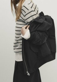 Schwarze Bomberjacke mit Reißverschluss, getragen über einem gestreiften weiß-grauen Pullover. Darunter ist ein weißes Hemd geschichtet, das weiche Texturen zeigt.