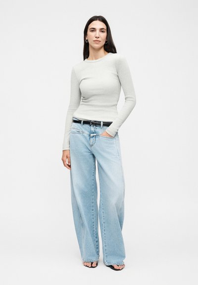 Γυναίκα με μακριά σκούρα μαλλιά που φοράει εφαρμοστή ανοιχτό γκρι μπλούζα με μακριά μανίκια, φαρδιά ανοιχτομπλε τζιν, μαύρη ζώνη και μαύρα παπούτσια με ανοιχτά δάχτυλα.