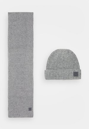 Lysegråt strikket uld tørklæde og matchende beanie-hat med foldet kant, begge med små mørke kvadratiske mærkelapper.