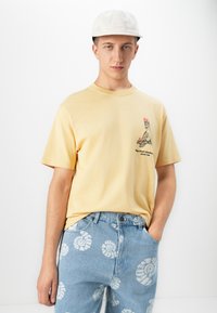 Ung mand med en beige kasket, en gul T-shirt med et flamingo-tryk og lyseblå jeans med hvide spiral-skaldmønstre.