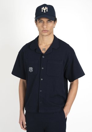 Jeune homme en chemise bleu marine à manches courtes boutonnée et casquette assortie avec des logos blancs, debout avec une main dans la poche sur un fond uni.