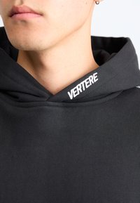Svart hettegenser laget av et teksturert stoff med en buet halslinje; har hvit brodert tekst "VERTERE" på kanten av hetten.