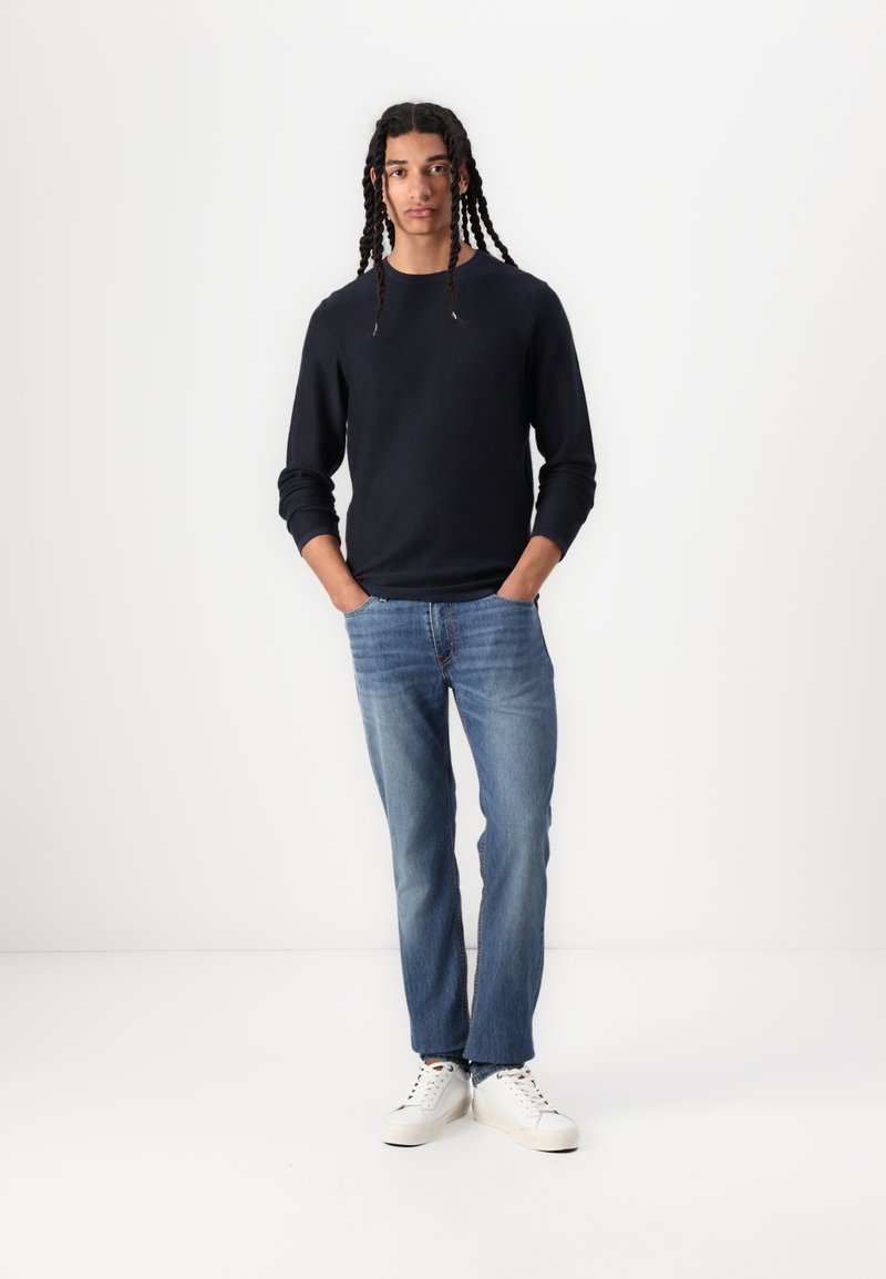 Junger Mann mit langen Zöpfen steht mit den Händen in den Taschen, trägt ein schwarzes Langarmshirt, blaue Jeans und weiße Sneakers vor einem weißen Hintergrund.