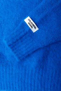 Material tricotat moale și pufoasă de culoare albastră strălucitoare, având pe margine o etichetă rectangulară albă cu inscripția "COLOURFUL REBEL".