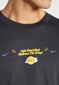 Juodas medvilninis marškinėlis su violetiniais ir geltonais grafikos užrašais "Ramink savo mintis, priimk sunkumą" su smegenų ikonėlėmis ir Lakers logotipu.