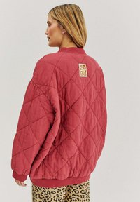 Červená prešívaná bunda s diamantovým vzorom, na zadnej strane s štvorcovou etiketou a grafikou preclíkov. Hladké manžety a lem.